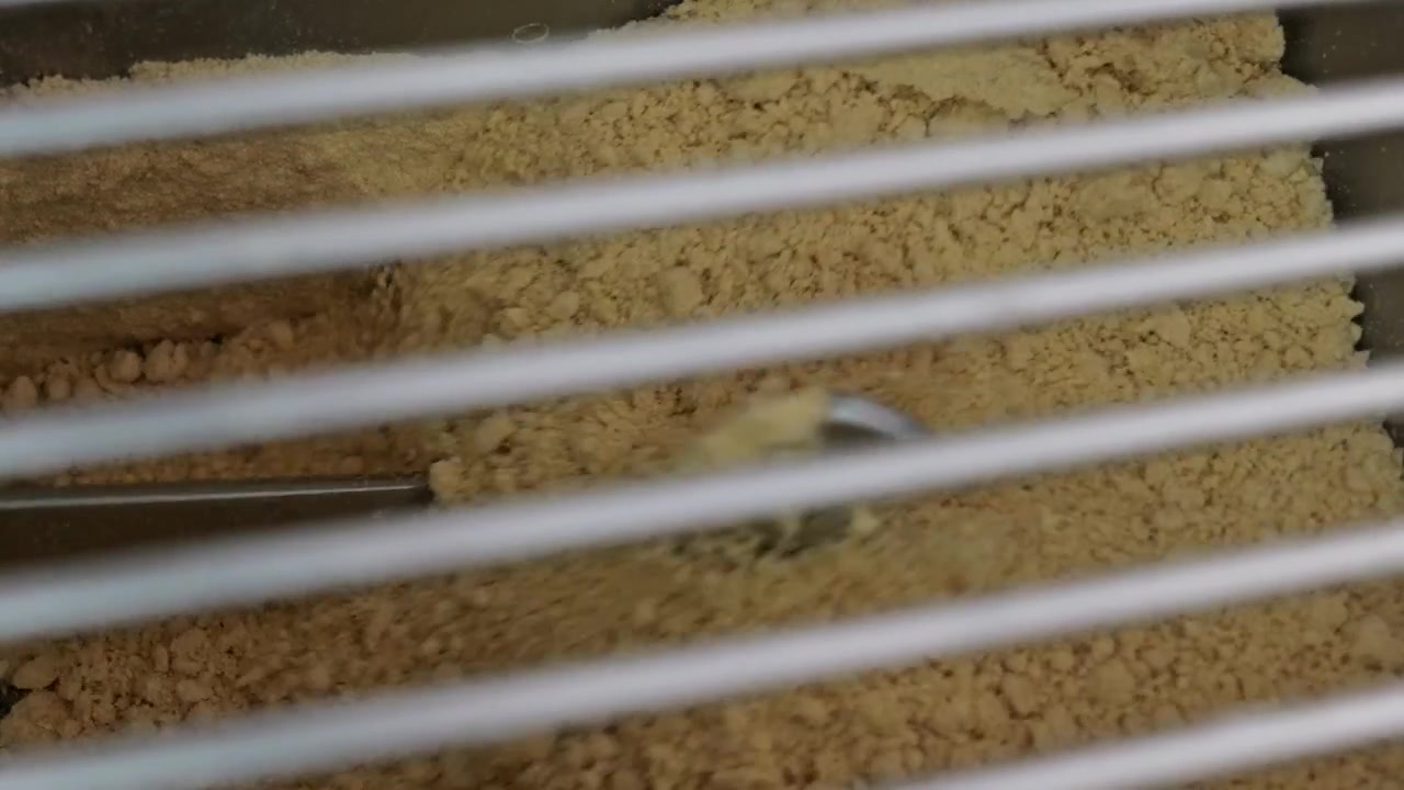 Low-angle close-up filmed through a protective safety grid as industrial mixing paddles blend dry pasta ingredients inside a commercial mixer