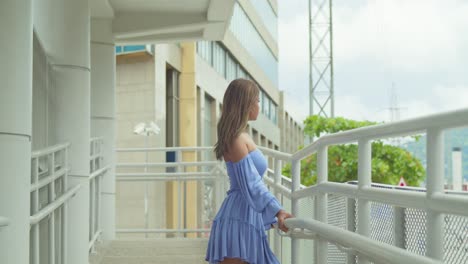 right pan while a young woman in a blue dress plays in her hair with city buildings in the background