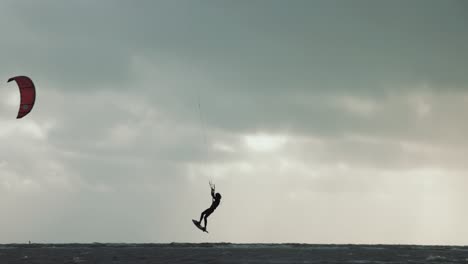 Kitesurfer-jumping-out-of-water-during-stormy-conditions,-while-the-sun-beams-through-the-darf-clouds-on-the-horizon