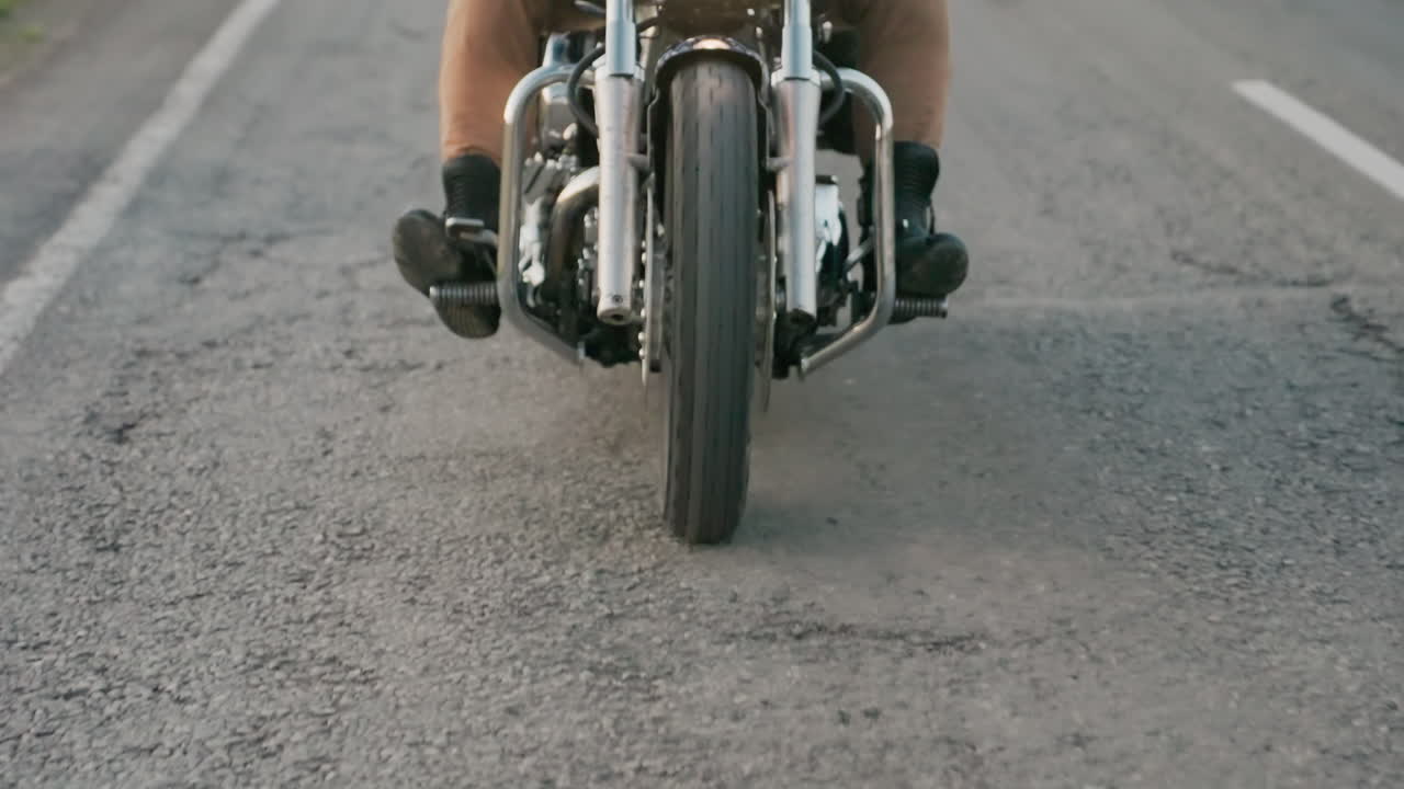 Vista della sezione inferiore di un motociclista che viaggia su una strada asfaltata, che mostra un primo piano della ruota anteriore, del telaio in metallo e degli stivali da motociclista che premono i pedali mentre il veicolo si muove costantemente lungo la campagna