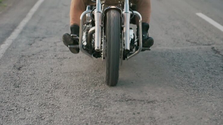 Vista della sezione inferiore di un motociclista che viaggia su una strada asfaltata, che mostra un primo piano della ruota anteriore, del telaio in metallo e degli stivali da motociclista che premono i pedali mentre il veicolo si muove costantemente lungo la campagna