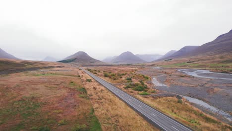 Aérea:-Revelación-De-Un-SUV-Blanco-Viajando-A-Lo-Largo-De-La-Carretera-De-Circunvalación-De-Islandia,-Que-Es-Una-Carretera-Panorámica-A-Través-De-Una-Pintoresca-Y-Remota-Zona-De-Fiordos-Que-Conduce-A-Picos-Gemelos-Con-Niebla-Y-Neblina-En-La-Distancia.