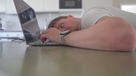 A young man puts his head down on the kitchen counter while looking up at his laptop computer