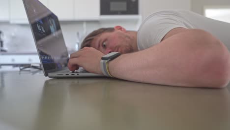 a young man puts his head down on the kitchen counter while looking up at his laptop computer