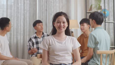 close up of a girl in plaid shirt smiling to camera. asian teen group sitting in chairs forming a circle discussing at home