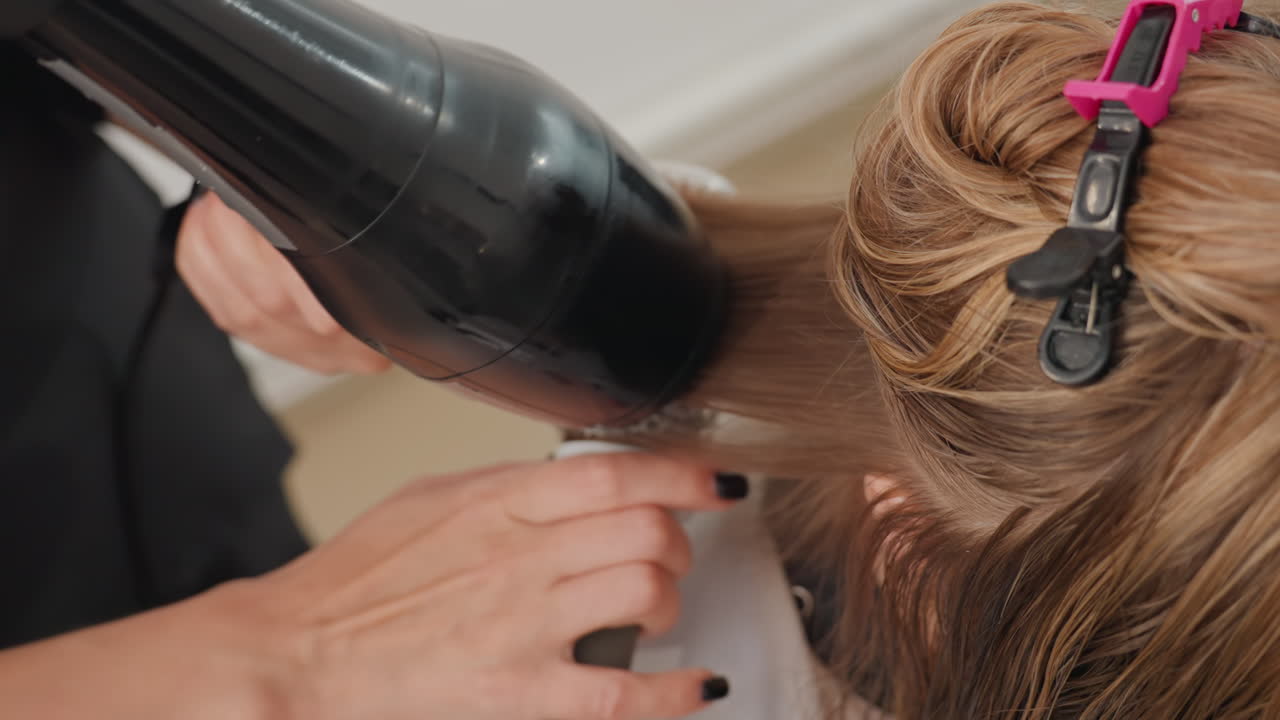 blonde hair sectioned and sprayed by stylist, closeup of hand applying product and arranging pink clip while directing dryer nozzle at roots, careful combing and tensioning for smooth base, clinical