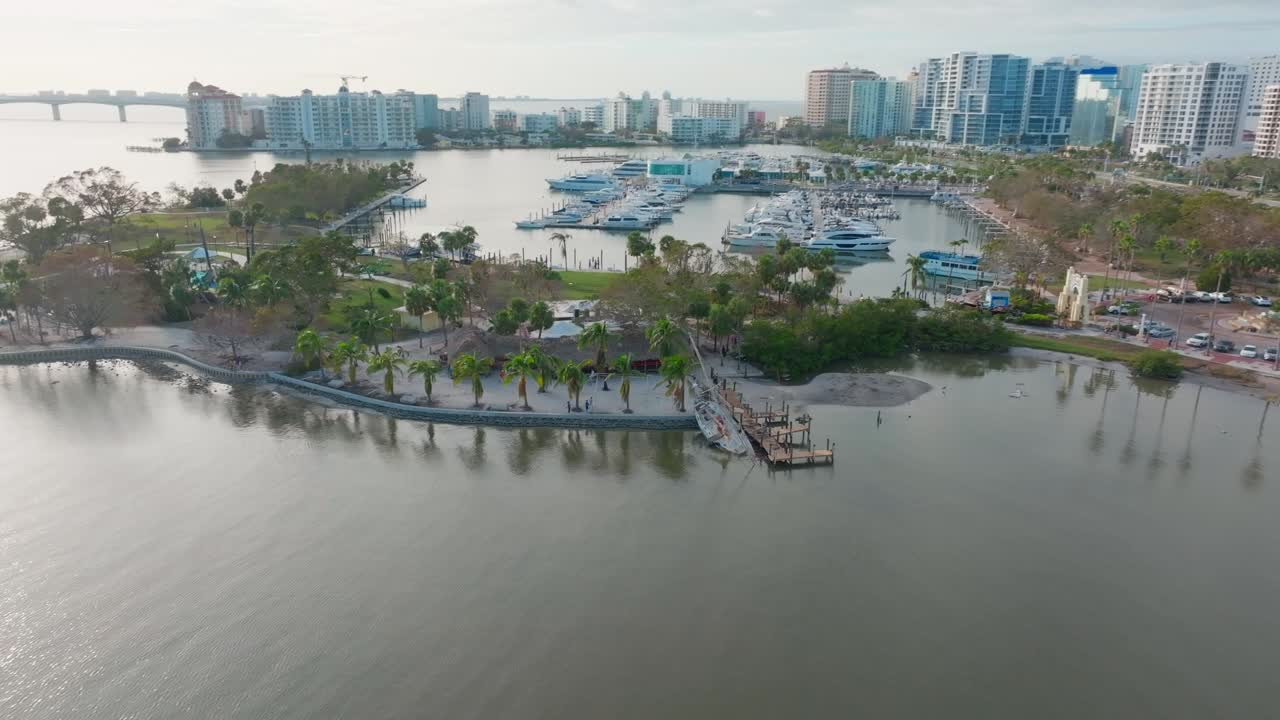Aerial bird’s-eye view over wrecked sailboat grounded in Bayfront Park, bow submerged. Pullback reveals Marina Jack and downtown Sarasota skyline in the background.
