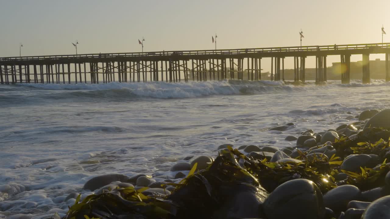 Gentle waves crash against a seaweed-covered rocky shore with a wooden pier in the background during golden hour