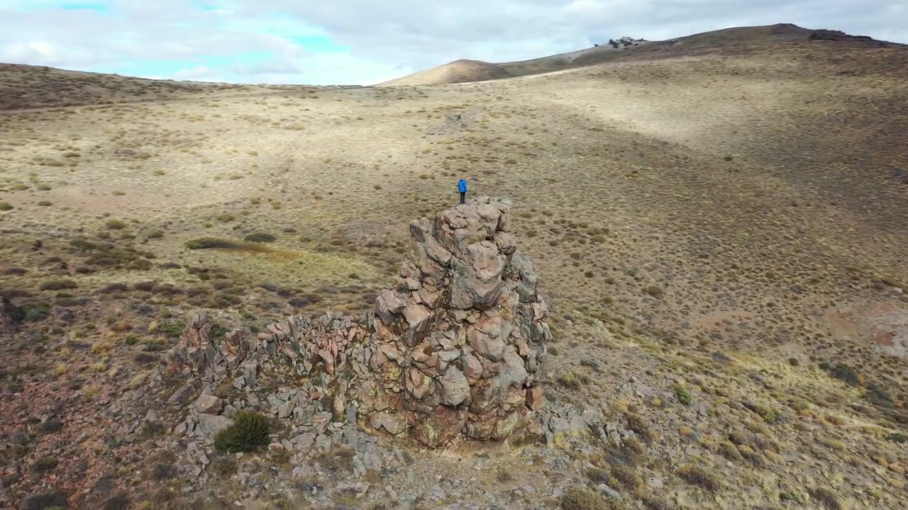 Una toma amplia y en picado de un excursionista solitario con chaqueta azul de pie en la cima de un pico rocoso en Llao Llao, Bariloche, Argentina. Destaca la escarpada y vasta naturaleza montañosa bajo cielos nublados.