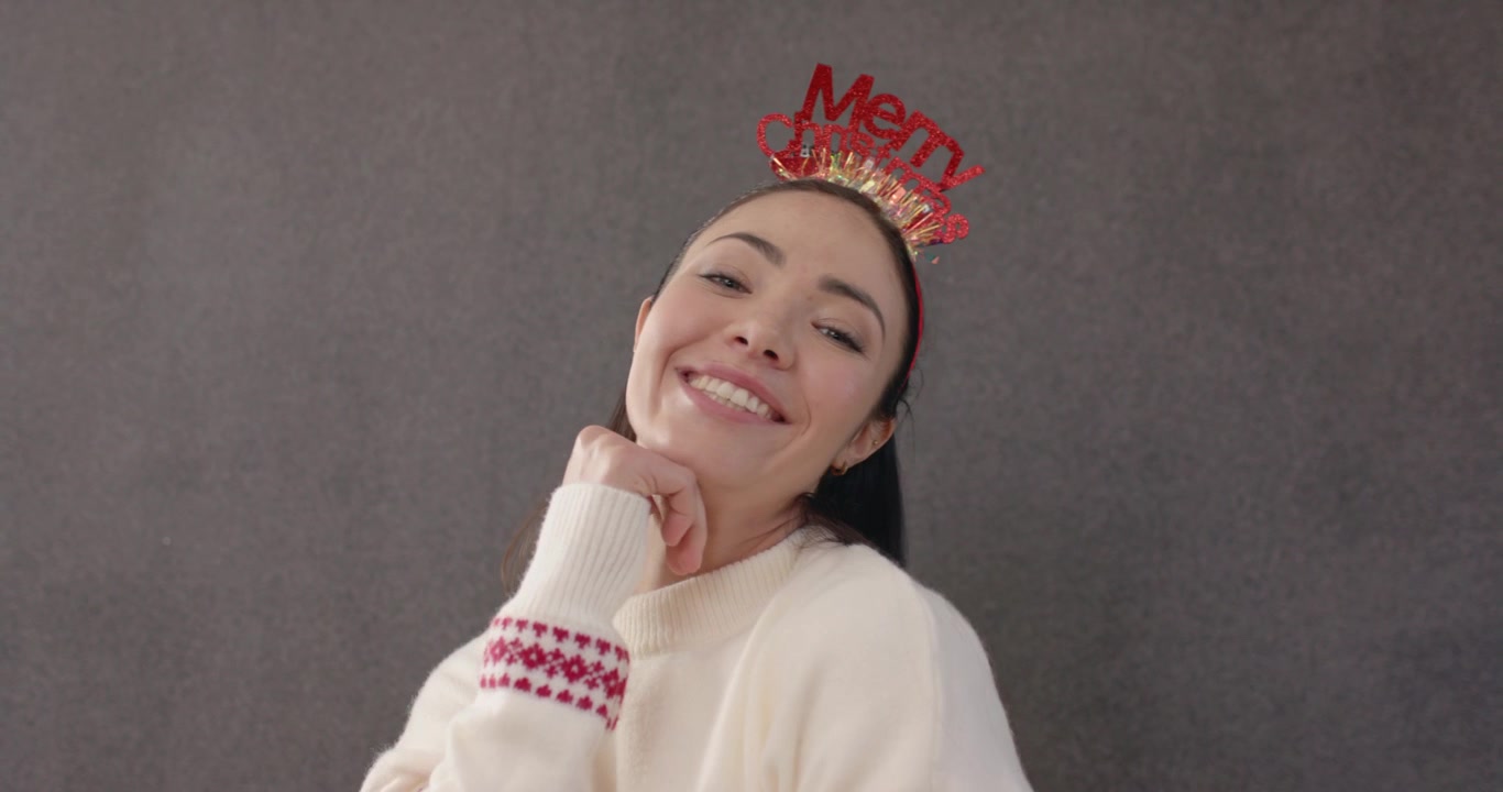 Người phụ nữ chỉnh sửa băng đô "Merry", tạo dáng, chỉ tay và nháy mắt vào máy ảnh trong studio, mặc áo len cho kỳ nghỉ lễ.