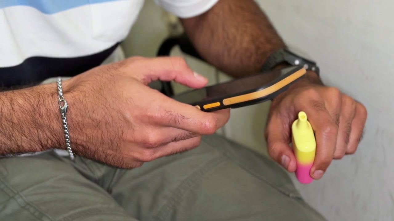 Primo piano delle mani di un uomo che scorre su uno smartphone nero mentre tiene una sigaretta elettronica usa e getta giallo brillante e rosa. La persona è seduta al chiuso, indossa abiti casual e controlla il telefono durante una pausa