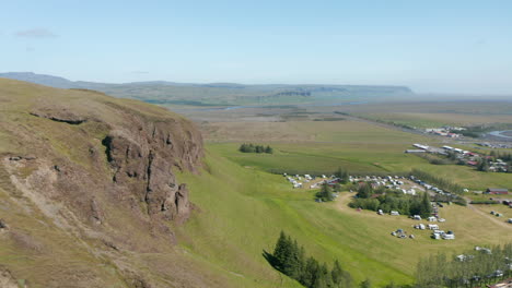 Drone-view-of-Iceland-grassy-green-highland-countryside-directly-above-a-small-town-near-river.-Birds-eye-of-touristic-campsite-near-a-small-village-in-lush-icelandic-highland