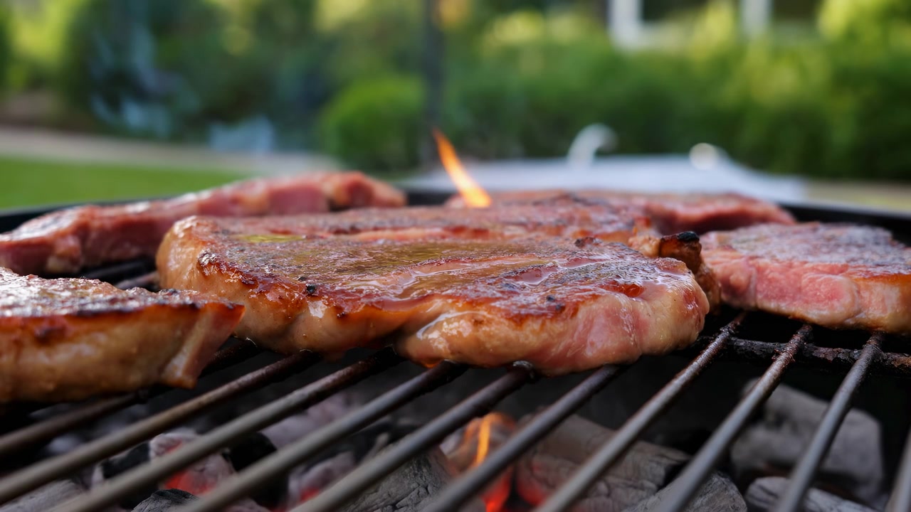 Close low-angle view shows browned meat cooking across hot barbecue grate, with glistening surfaces, orange flame bursts below, and light smoke drifting upward in an outdoor setting.