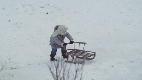 young girl slowly pushes wooden sled up a snow covered hill, wide shot