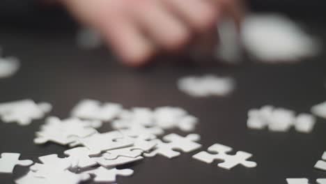 Close-up-slow-motion-of-white-puzzle-pieces-laying-randomly-on-a-dark-table-with-a-hand-taking-a-piece