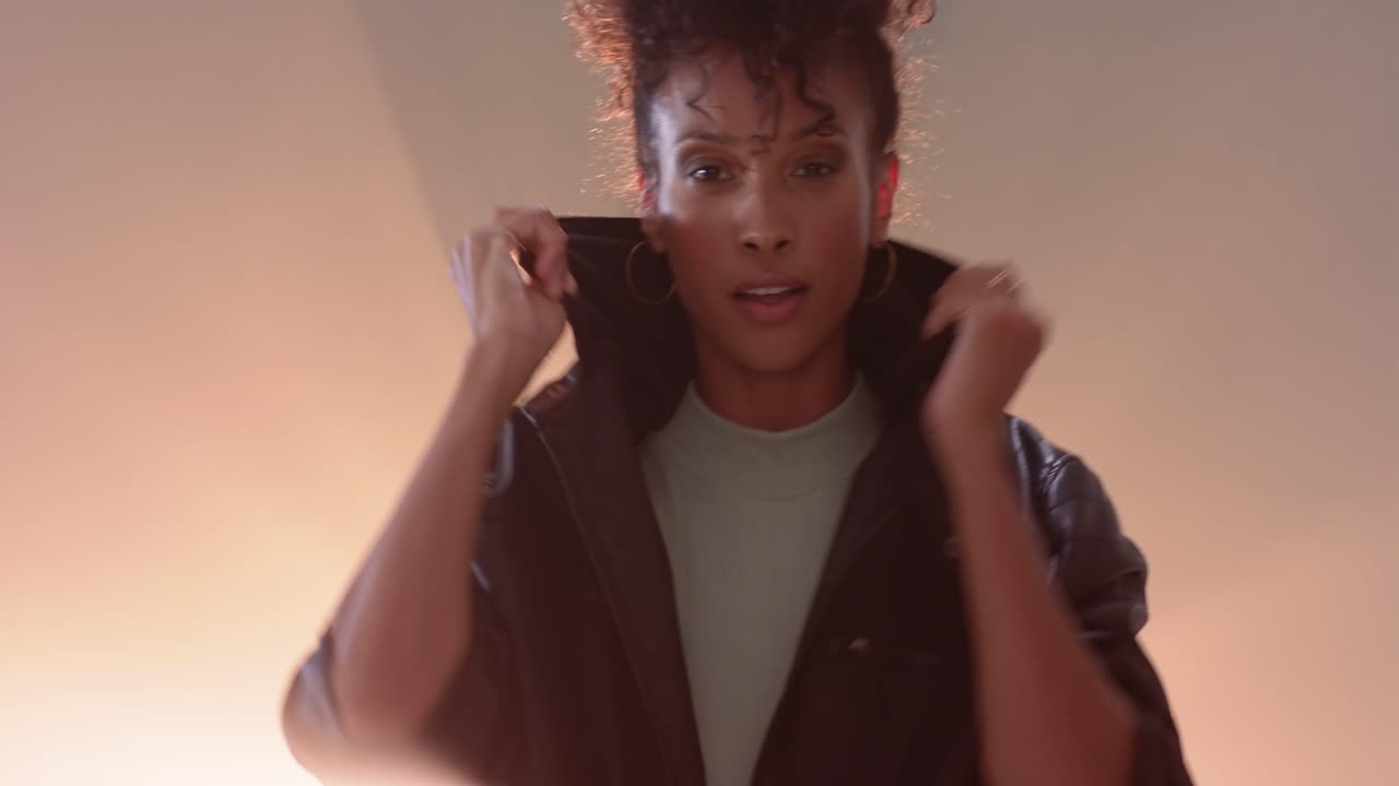På anvisning lader en afroamerikansk kvinde i 20'erne en mørk jakke glide ned, mens hun modellerer i studie med modlys.