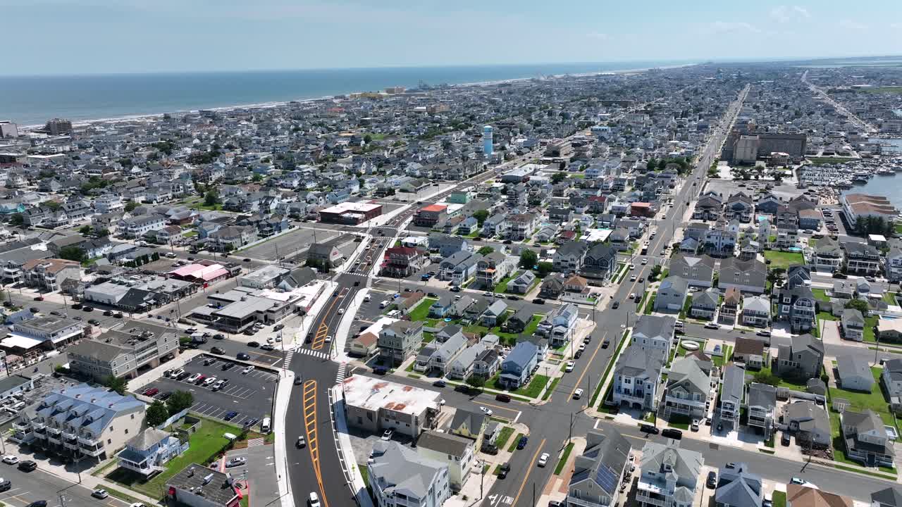 Aerial rising wide shot showing Wildwood City in New Jersey with Atlantic ocean in background. Sunny summer day with cars on main street in neighborhood. Beach houses and apartment blocks