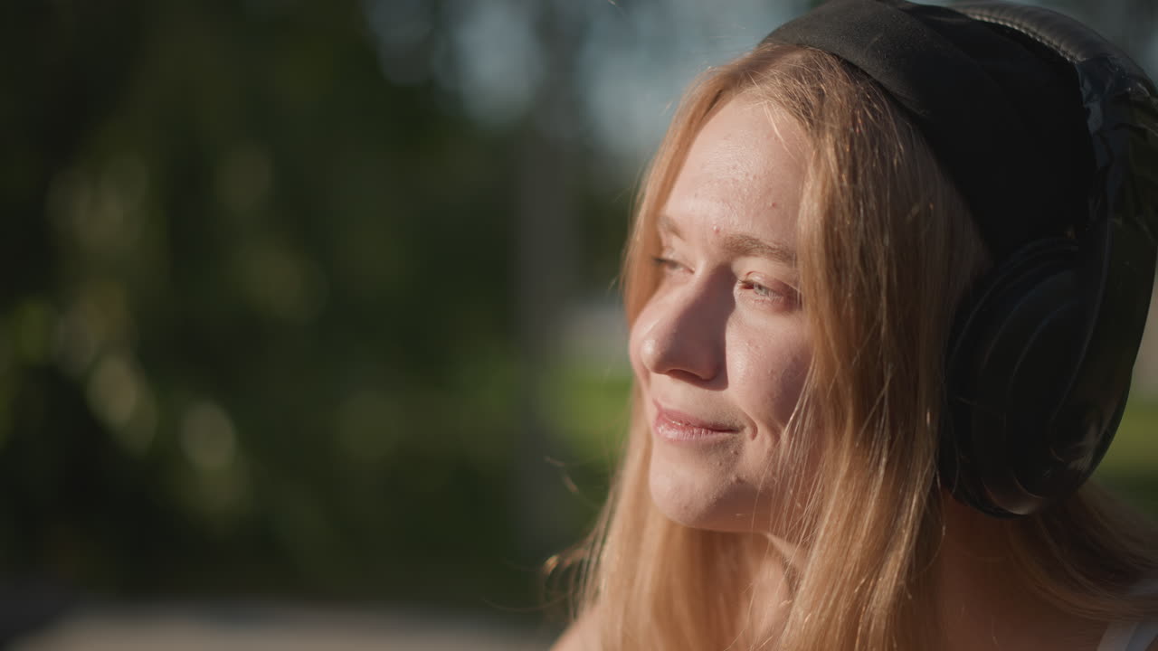 Mulher branca jovem ouvindo música com fones de ouvido grandes, tomando shake de proteína em uma garrafa de shaker transparente em um parque ensolarado, olhos fechados, sorriso relaxado, levemente balançando a cabeça, luz dourada, verão