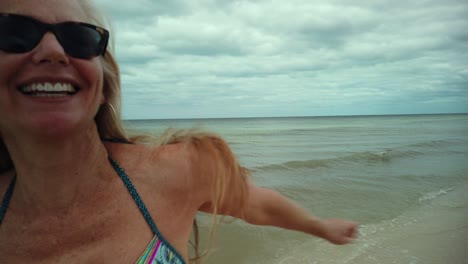 el primer plano de una mujer madura riendo y juguetona en bikini y gafas de sol se da la vuelta y corre en la playa
