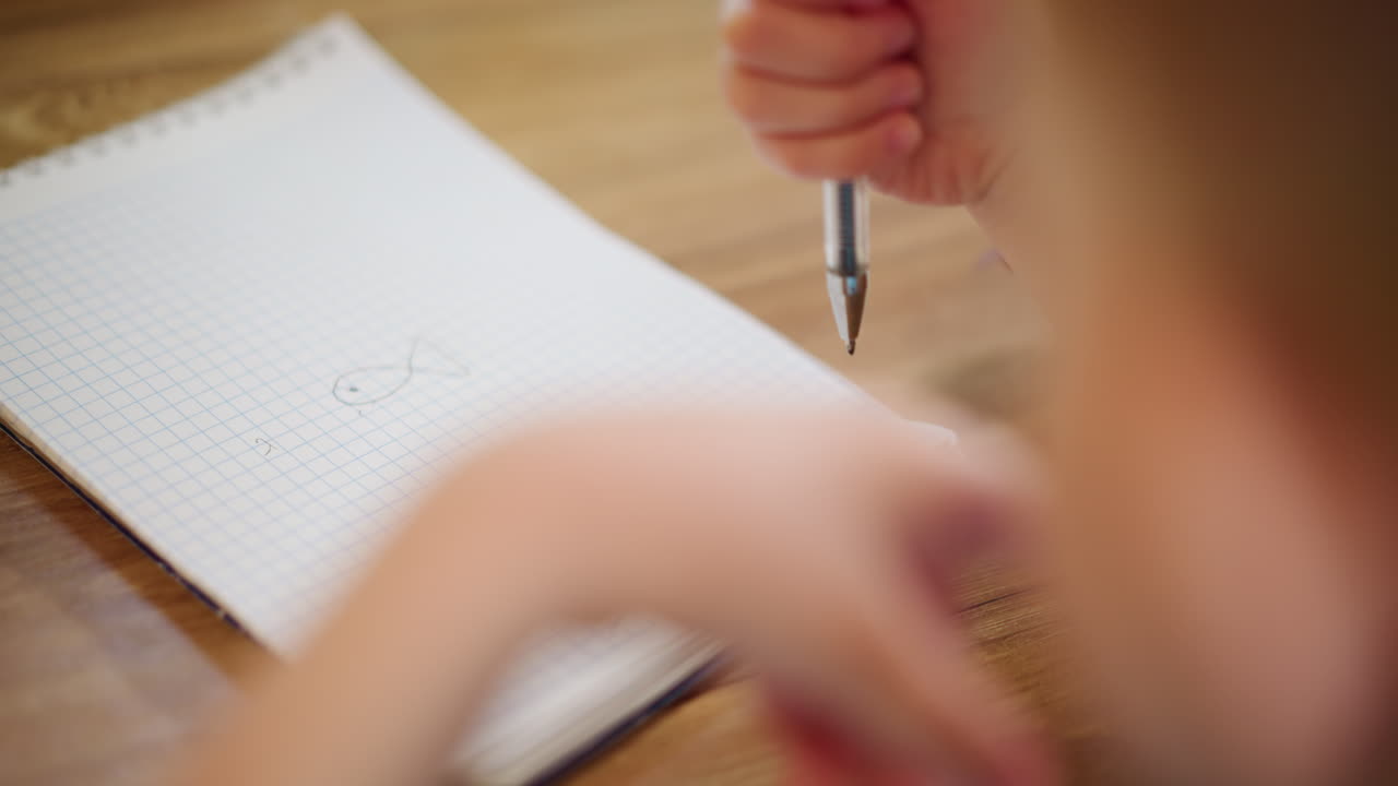 Child hand holds pen while practicing drawing shapes on squared notebook page during learning activity at home, showing early writing skills, concentration, and creativity with closeup view