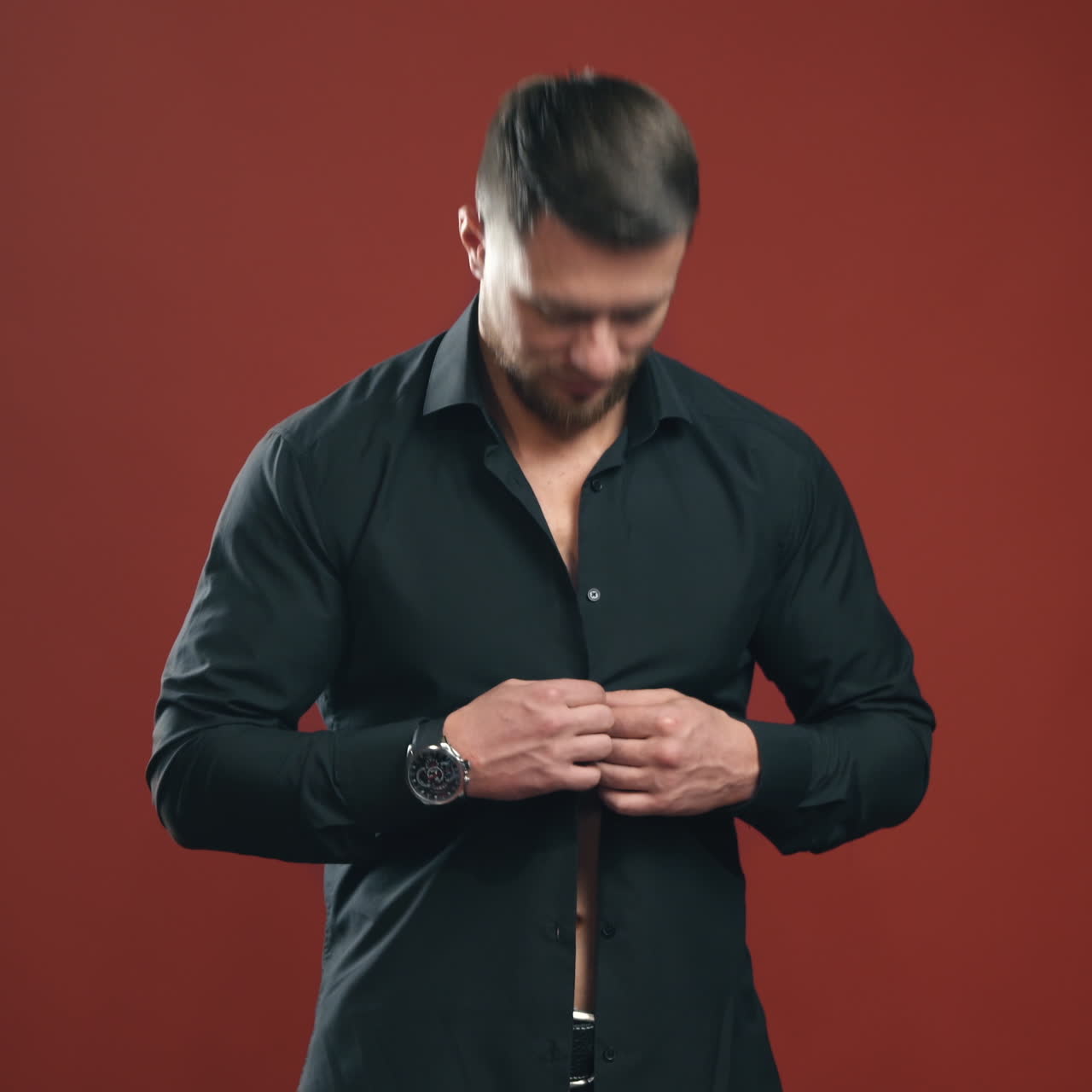 Sexy man in black shirt. Handsome male unbuttons his shirt and shows his naked torso on dark red studio background. Bearded man with sporty body indoors.