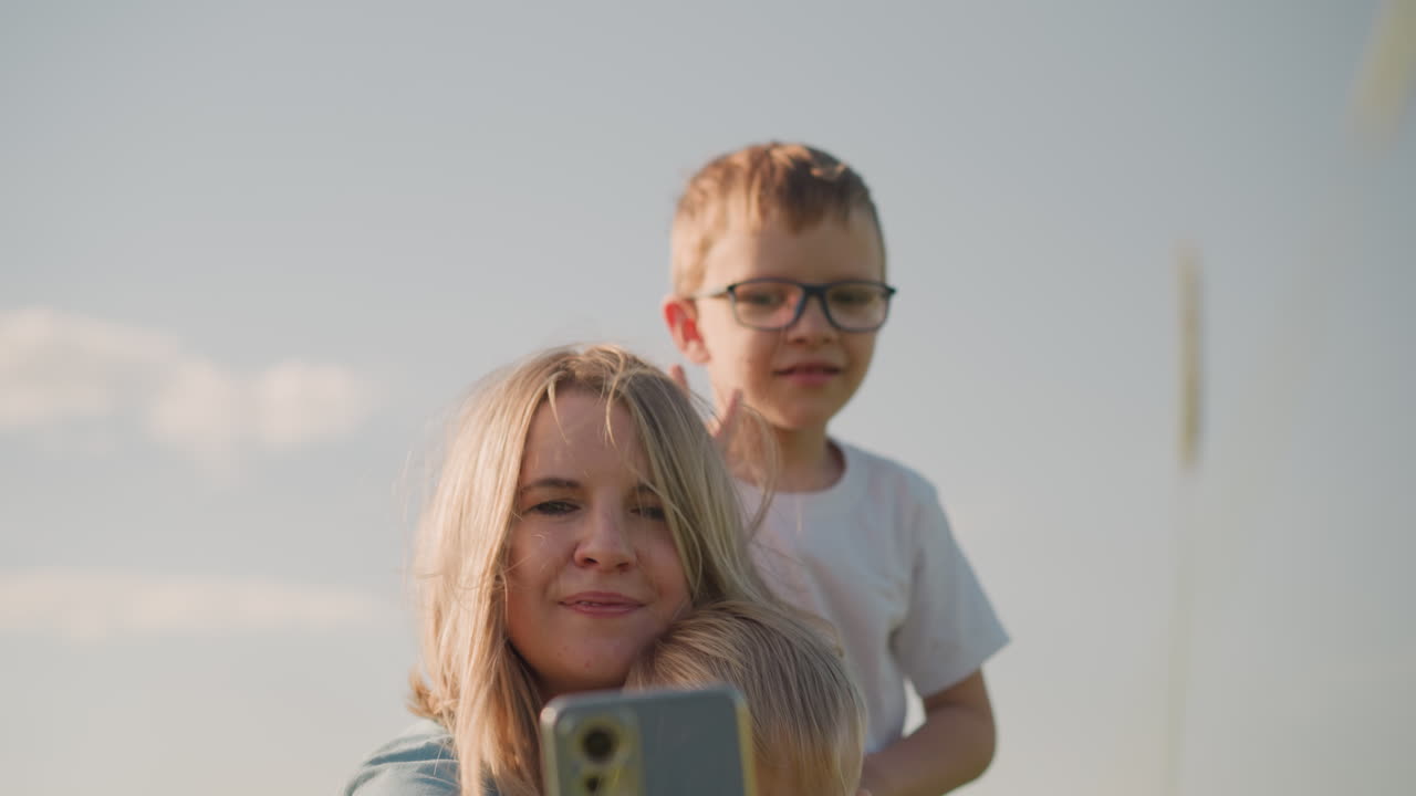 A woman sitting on the grass, takes a picture with her smartphone. Her son, wearing glasses, is behind her making a playful gesture to appear in the picture