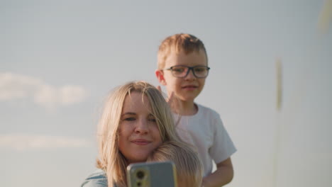 a woman sitting on the grass, takes a picture with her smartphone. her son, wearing glasses, is behind her making a playful gesture to appear in the picture