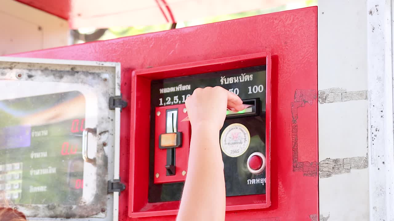 A person inserts cash into a red coin-operated fuel dispenser under bright daylight at Khao Rang Viewpoint, Phuket