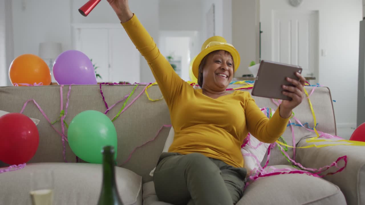 Happy african american senior woman using party blower making new year's eve tablet video call
