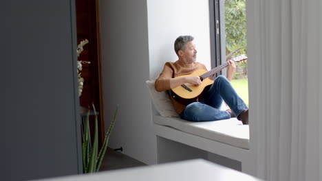 Hombre-Birracial-Senior-Enfocado-Sentado-En-El-Asiento-De-La-Ventana-Tocando-La-Guitarra-En-Casa,-Espacio-Para-Copiar,-Cámara-Lenta