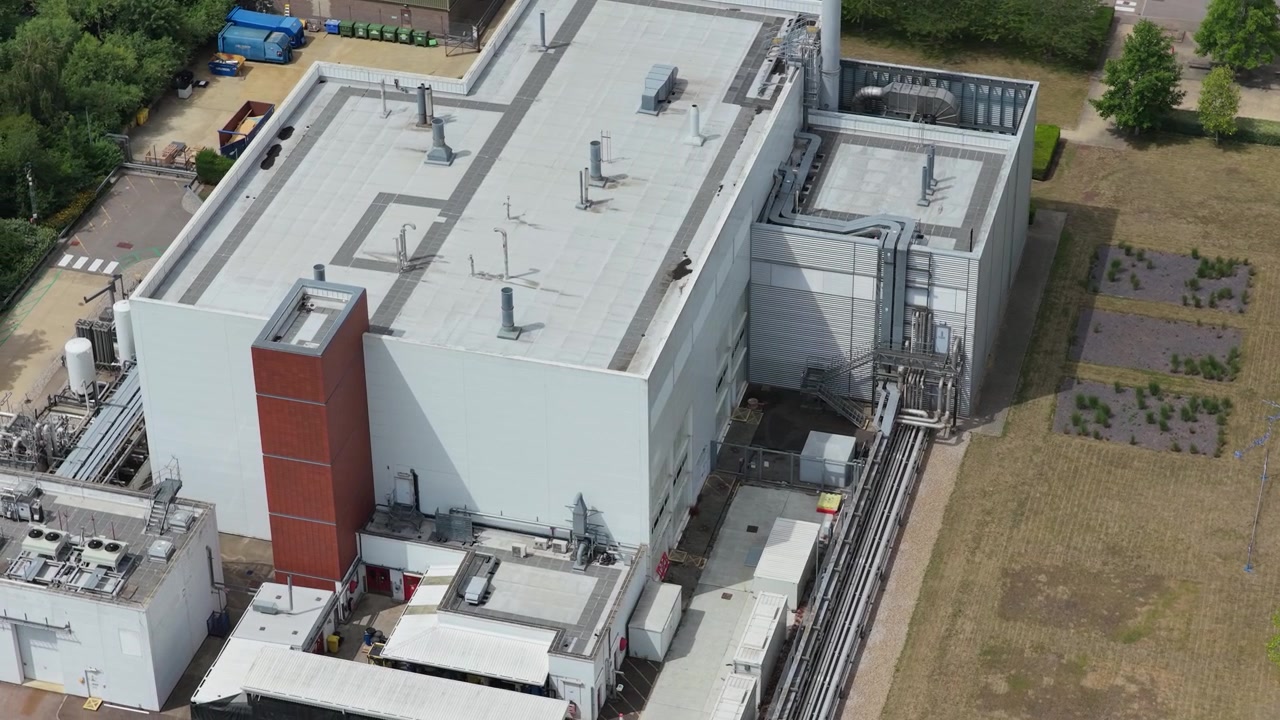 Gran instalación industrial con un edificio blanco y detalles marrones, rodeado de árboles verdes y tuberías en una tarde soleada en una zona rural a mediodía.