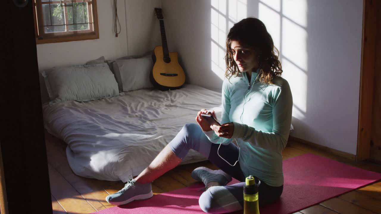 mujer de raza mixta feliz preparándose para el entrenamiento, usando un teléfono inteligente sentada en el suelo en un dormitorio soleado