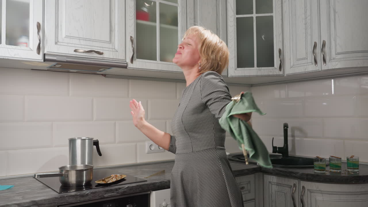Graceful lady with short bob hair in long gown wipes kitchen utensil with towel while emotionally raising hands to music, surrounded by oven, cabinets, and glass cups filled with coconut wax