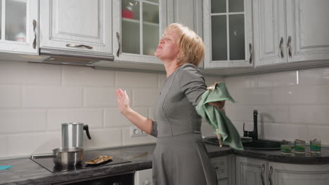 graceful lady with short bob hair in long gown wipes kitchen utensil with towel while emotionally raising hands to music, surrounded by oven, cabinets, and glass cups filled with coconut wax