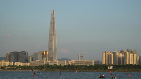 Grupo-De-Personas-Practicando-Windsurf-En-El-Río-Han-Junto-A-La-Famosa-Torre-Mundial-Lotte-De-Corea-En-La-Hora-Dorada---Espacio-De-Copia---Jamsil,-Tukseom-Park-Surf-Club