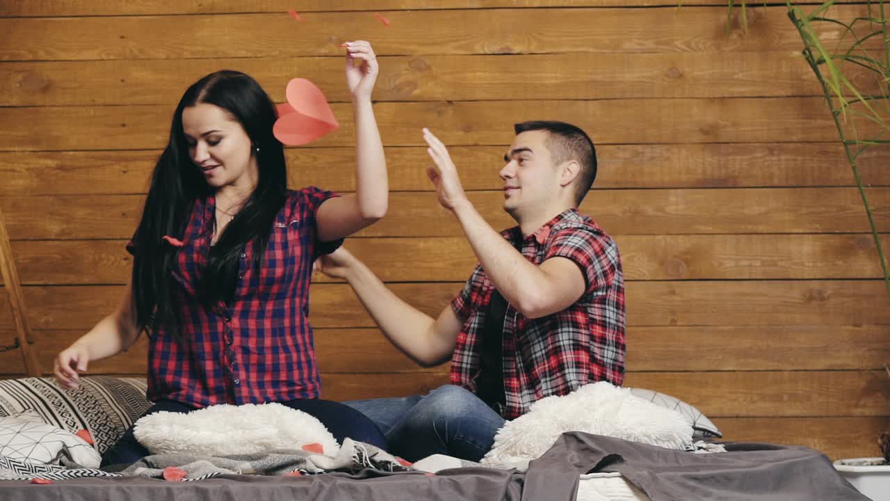 enamored man and woman are sitting on the bed and throwing up red paper hearts with their hands on Valentine's Day