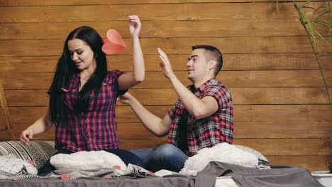 enamored man and woman are sitting on the bed and throwing up red paper hearts with their hands on valentine's day
