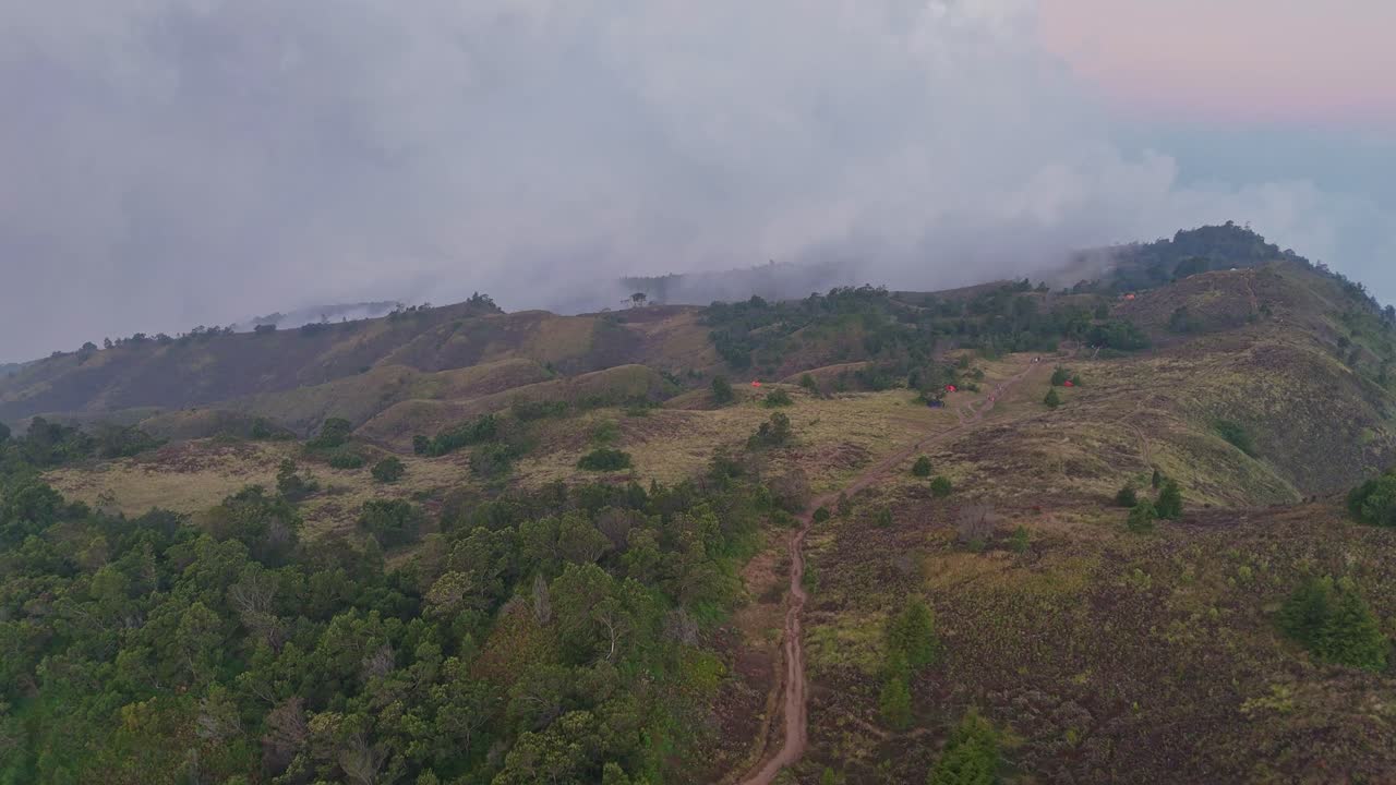 Aerial view of mountain ridge covered in grassland and scattered trees with narrow hiking trail winding through the landscape. Misty clouds roll over the hills in the background. Mount Prau, Indonesia