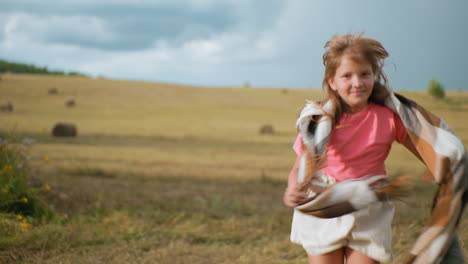 smiling little girl in pink top and shorts joyfully running across vast open field with blanket draped over shoulders, wind blowing through hair, golden hay bales scattered in background