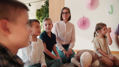 Una-Chica-Rubia-Con-Un-Peinado-Bob-Con-Gafas-Y-Una-Camisa-Blanca-Y-Un-Hombre-Con-Una-Camisa-Violeta-Están-Sentados-En-Un-Círculo-Con-Niños-En-El-Suelo-Y-Hablando-En-Su-Lección-Introductoria-Sobre-Cómo-Prepararse-Para-La-Escuela.