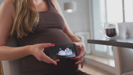 close up of pregnant woman at home holding mobile phone with ultrasound scan of baby