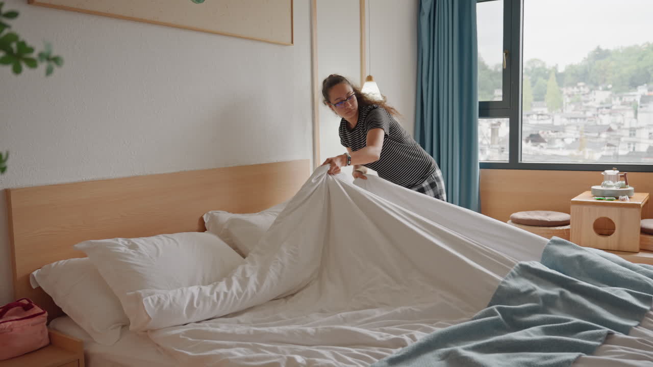 Viajero caucásico arregla la cama del hotel, tira del edredón, alisa la ropa de cama, acomoda las almohadas, verifica la pulcritud de la habitación; movimiento tranquilo y eficiente, vista de la ciudad por la ventana, la luz del día llena la habitación, escena de hospitalidad moderna.
