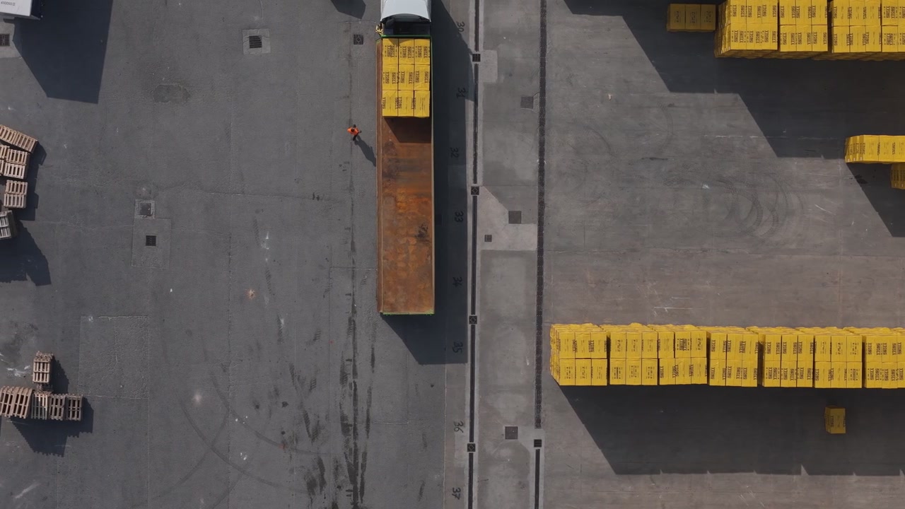 Top down aerial view of industrial yard with truck loading yellow construction blocks for export logistics, showing cargo handling and manufacturing distribution activity, drone lateral slide shot.