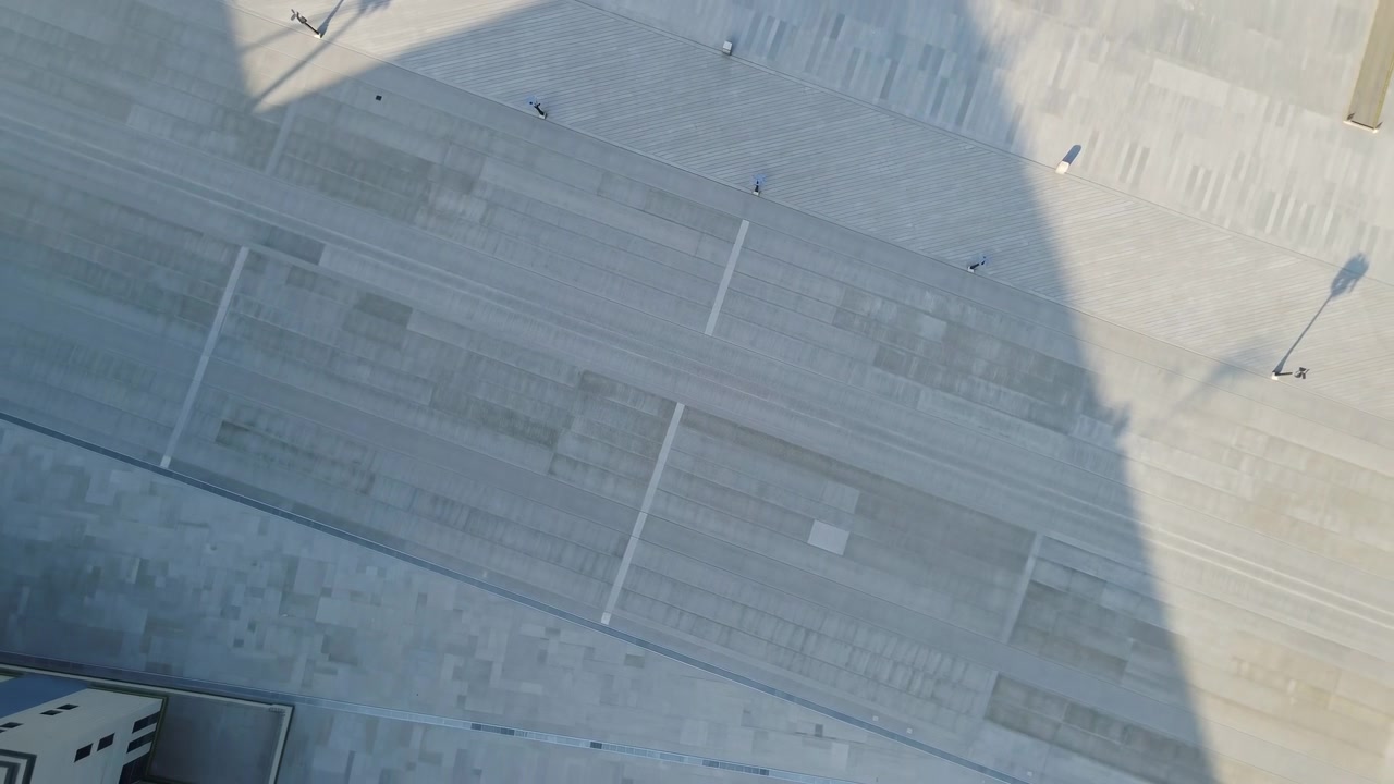 Barrido progresivo de una gran sombra triangular cruza una plaza de hormigón moderna y vacía y terrazas escalonadas, mientras la luz solar de ángulo bajo desde la parte superior izquierda produce reflejos azulados fríos y un fuerte contraste.