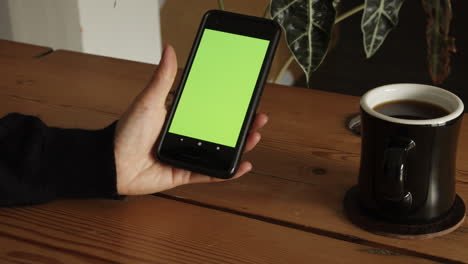 Female-hand-sitting-on-table-drinking-coffee-and-swiping-up-on-green-screen-of-smartphone