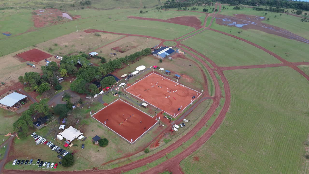 A red sand arena set in a scenic landscape hosts an equestrian sports event outdoors.