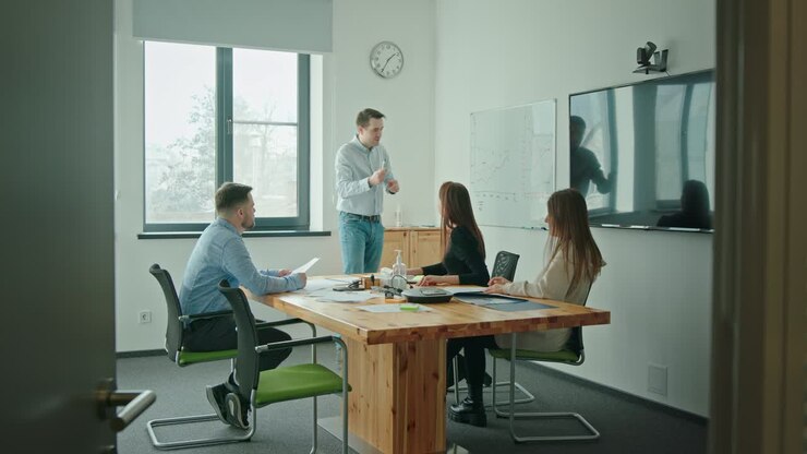 Un team di giovani che lavorano insieme in un'azienda in un ufficio moderno, seduti a un tavolo di negoziazione, discutono un nuovo progetto mentre un uomo disegna un grafico su una lavagna bianca