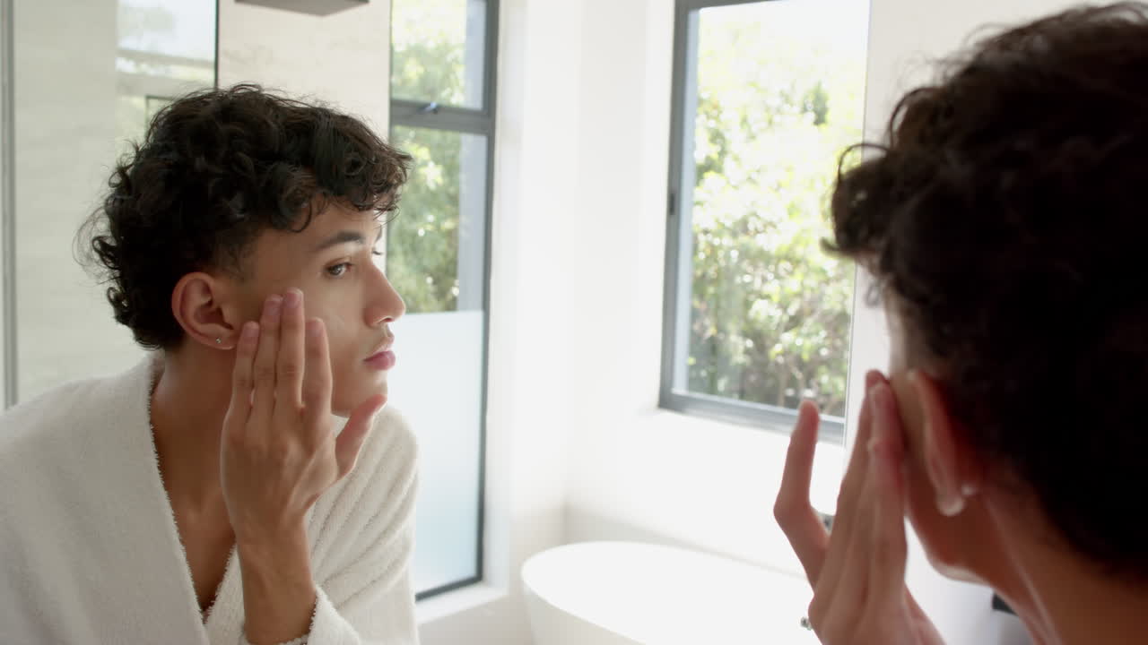 Applying skincare, non binary person looking in mirror in bright bathroom