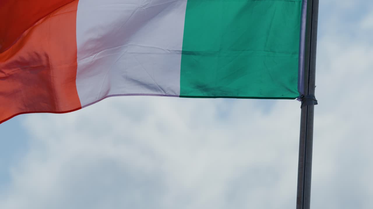 Una bandiera irlandese sventola dinamicamente su un pennone contro un cielo nuvoloso, luce diurna, telecamera fissa, colori vivaci