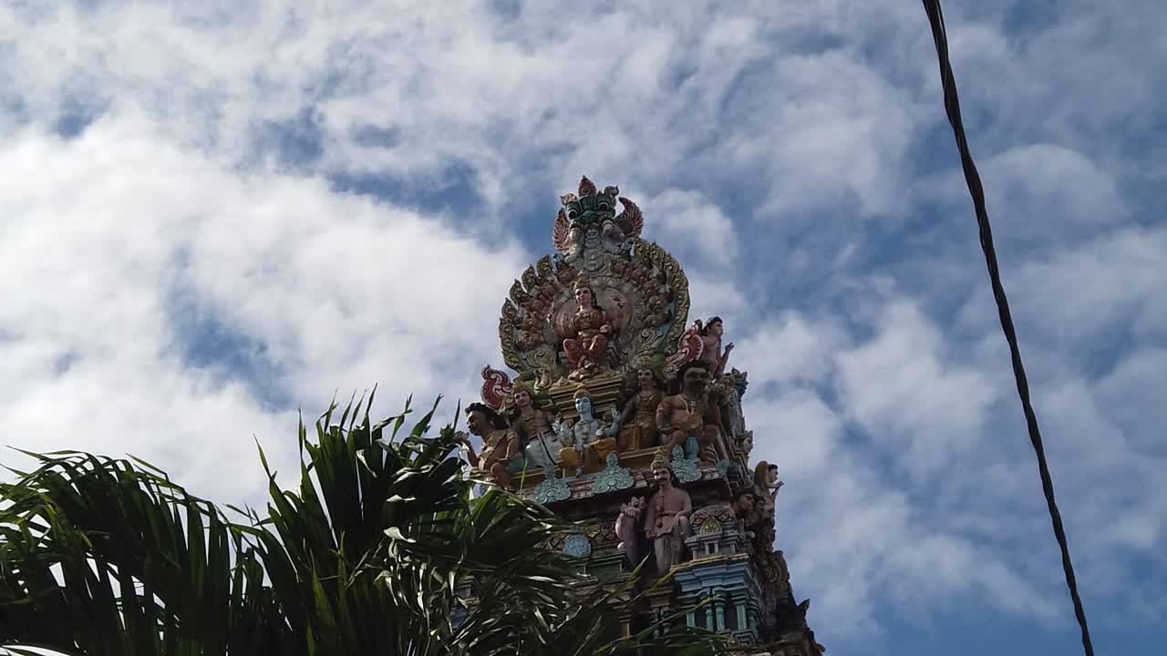 una toma de la torre de un templo hindú lleno de deidades caminando por la calle, con una palma en el medio y muchas nubes diminutas en el cielo